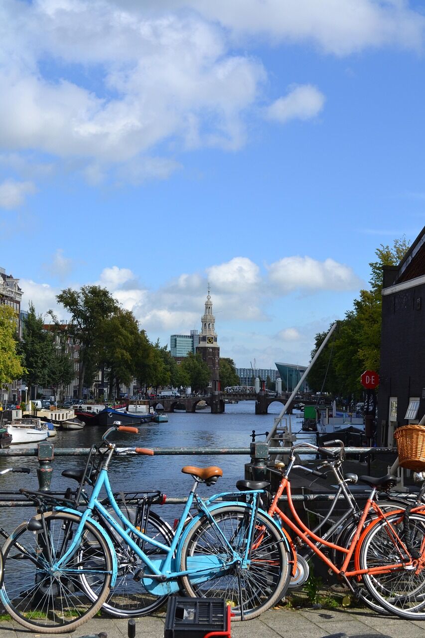 Bicycles parked by a canal in Amsterdam with a historic tower and bridge in the background.